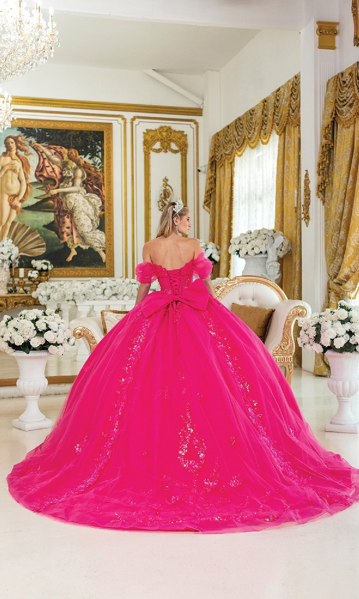 A woman in the Quinceanera Dress 5006 By Dancing Queen, a fuchsia gown with a corset bodice and large bow, stands in a luxurious room decorated with white flowers, gold accents, and classical artwork.